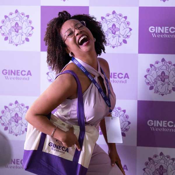 Participante sorrindo com a sacola oficial do Gineca Weekend em frente ao painel do evento.